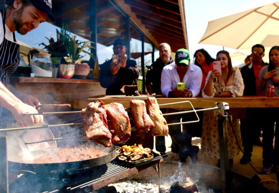 Cook turning cuts of meat on an open asado grill while travelers watch at a winery restaurant
