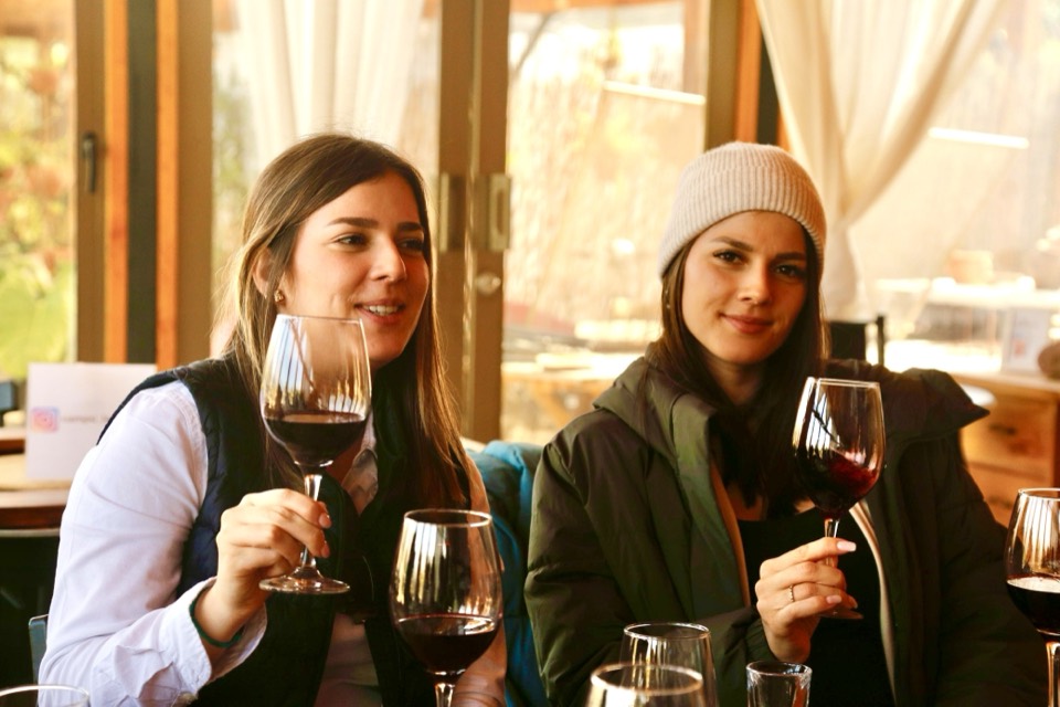 Two friends smiling and holding glasses of red wine at a winery